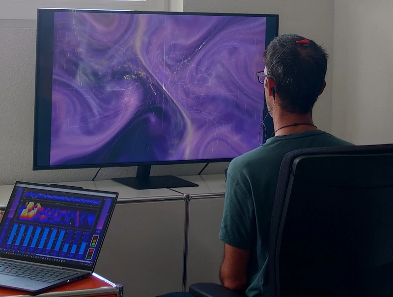 Personne faisant un entraînement de neurofeedback. Le patient suit sur l'écran un feedback basé su le signal des électrodes posées su sa tête. Le thérapeute peut simultanément observer en détails les signaux bruts sur son écran et adapter le feedback aux objectifs définis préalablement.